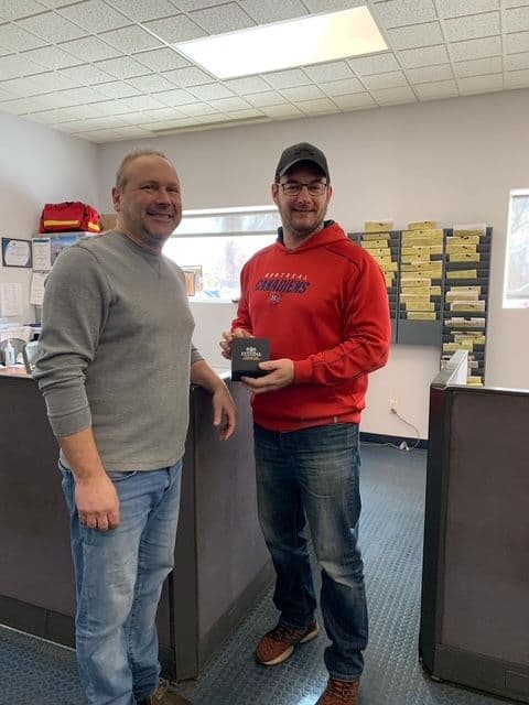 Two men standing in an office, one in a gray sweater and the other in a red sweatshirt holding a black trophy with a smile.