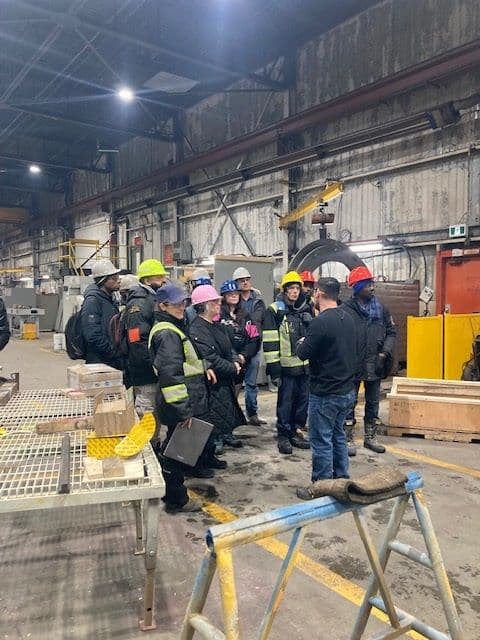 Workers in safety gear and colored helmets listen to a supervisor in a factory, surrounded by industrial equipment and metal tables.