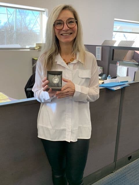 Smiling woman with blond hair and glasses, holding a box containing a silver watch, standing in an office.